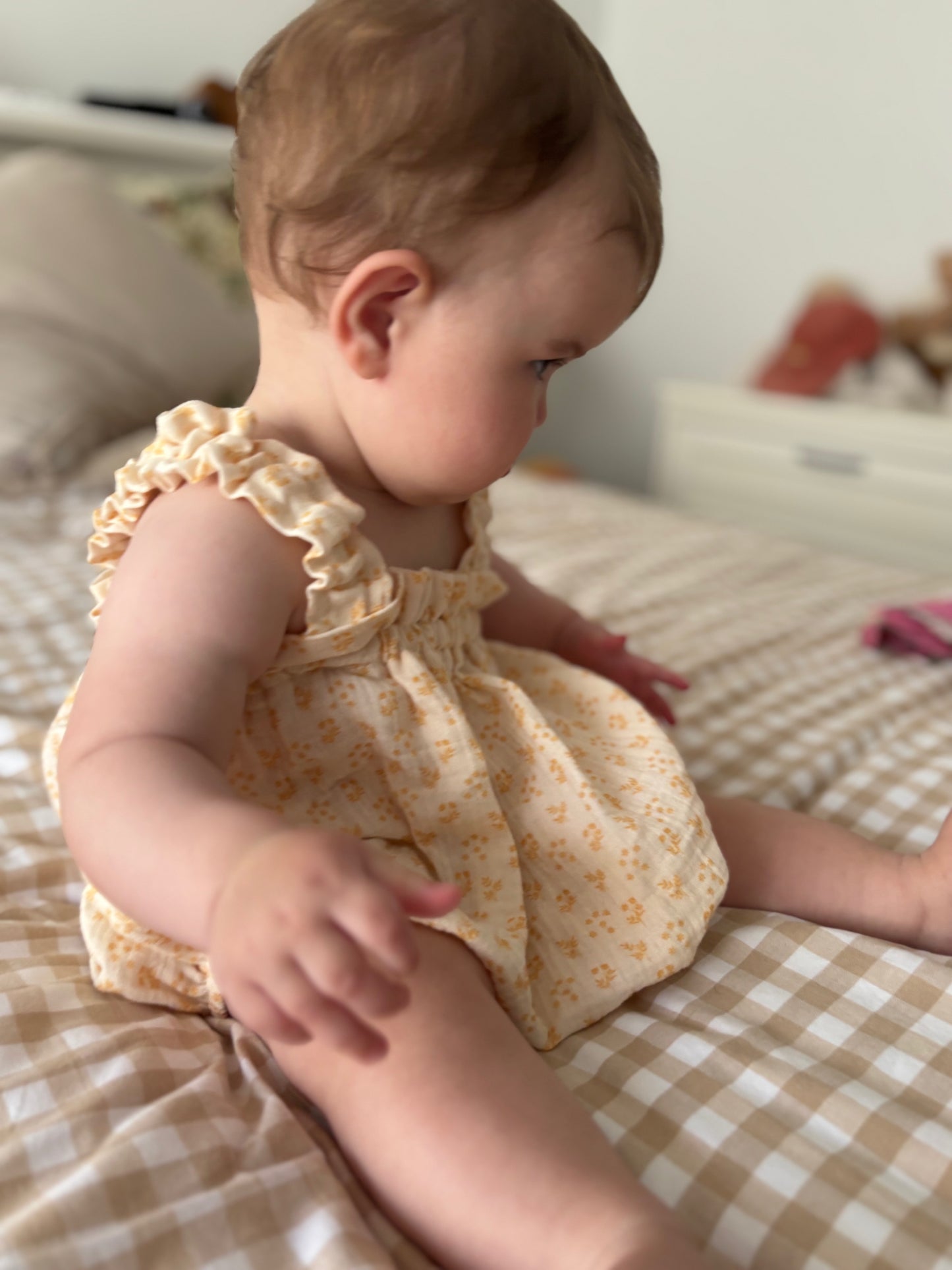Baby in a yellow floral outfit sitting on a checkered bed.
